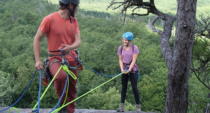 Two people wearing safety gear are secured by ropes near the edge of a cliff. One person appears to be an instructor, giving direction to the other person. There is a green wooded area below them.